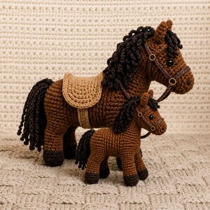 May include: Two crocheted horse figures, one large and one small, in shades of brown with black manes and tails. The larger horse has a tan saddle. Both have brown bridles with metal rings. The figures are set against a textured, neutral background.