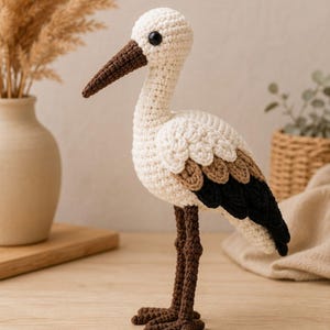 Puede incluir: Una figura de cigüeña tejida a ganchillo con cuerpo color crema, un pico largo marrón y detalles en las alas de color negro y marrón. El ave tiene un ojo negro redondo y se sostiene sobre dos patas largas y texturizadas de color marrón.