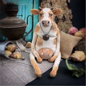以下が含まれることがあります： 白と茶色の斑点模様の手作りぬいぐるみ牛。 牛は首にベルを付けて座っています。 このぬいぐるみは、ユニークな手作りのアイテムです。