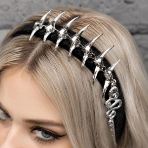 Puede incluir: Una diadema negra con detalles de garras y calaveras plateadas. Un dije de serpiente plateada cuelga de la diadema. El accesorio se usa sobre cabello rubio.