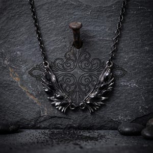 Puede incluir: Un collar oscuro de estilo g&oacute;tico con un par de alas estilizadas. Las alas son de metal oscuro y est&aacute;n adornadas con piedras preciosas negras. El collar cuelga de una cadena oscura, creando un contraste llamativo.