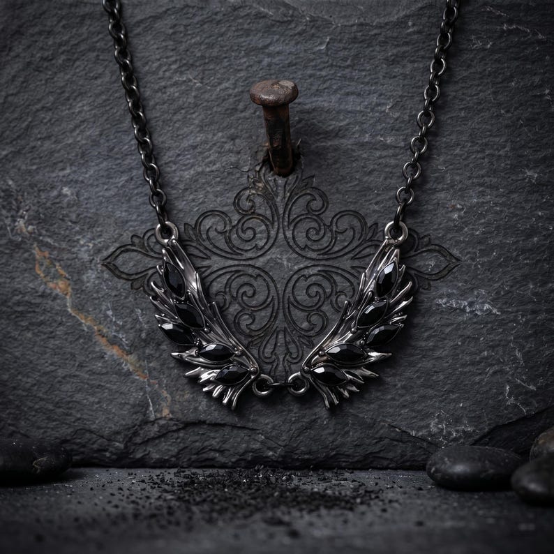Puede incluir: Un collar oscuro de estilo g&oacute;tico con un par de alas estilizadas. Las alas son de metal oscuro y est&aacute;n adornadas con piedras preciosas negras. El collar cuelga de una cadena oscura, creando un contraste llamativo.
