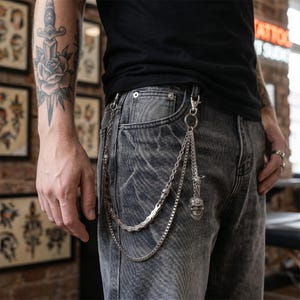 May include: Silver-toned chain wallet accessory with a skull pendant and a bike chain design. The chain is attached to the belt loop of gray denim jeans. The accessory adds a punk rock aesthetic.