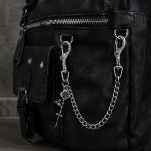 Puede incluir: Un bolso de cuero negro con una cadena de metal plateado sujeta por dos mosquetones grabados con motivos florales. Un pequeño dije de rosa plateada cuelga de la cadena, la cual está asegurada al bolsillo exterior y al cuerpo del bolso.