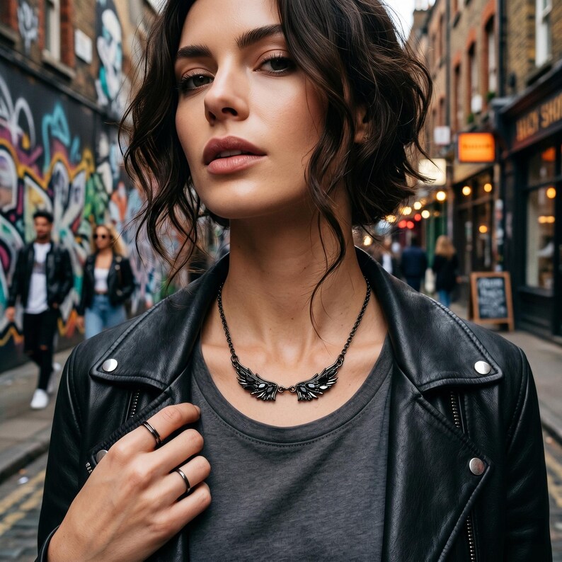 Puede incluir: Una mujer con una chaqueta de cuero negra y una camiseta gris, con un collar negro con forma de alas y dos anillos negros. El fondo muestra una pared cubierta de grafitis y una escena callejera.