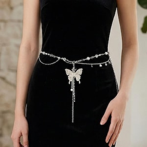 Puede incluir: Una cadena de cuerpo plateada con un colgante de mariposa. La cadena presenta pequeñas cuentas y un dije de mariposa colgante con una larga borla de cuentas. La joya se usa sobre un vestido negro.