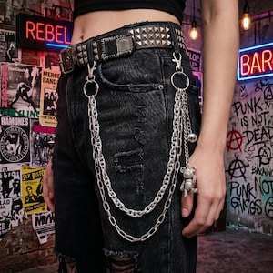 May include: A studded brown leather belt with silver chain pant accessories. The chains hang from a silver ring attached to the belt loops of black denim shorts. A small teddy bear charm and a silver ball dangle from the chains.