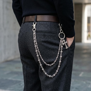 Peut inclure: Une chaîne de portefeuille argentée avec un pendentif en forme de croix et des chaînes de perles décoratives est suspendue à une ceinture en cuir marron. La chaîne est attachée à un pantalon gris.