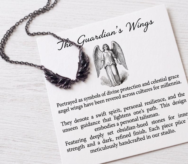 Puede incluir: Un collar oscuro, con un tono de metal de ca&ntilde;&oacute;n, con un colgante en forma de ala con piedras de color obsidiana. El collar se muestra con una tarjeta que dice "The Guardian's Wings" y describe el simbolismo de las alas de &aacute;ngel.