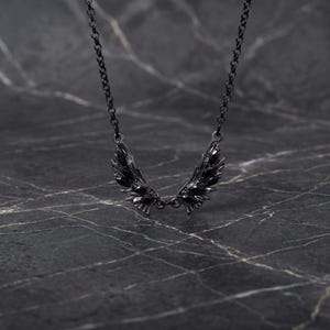 Puede incluir: Un collar negro con un colgante de alas estilizadas. Las alas, con detalles texturizados, cuelgan de una fina cadena. La joya destaca sobre un fondo oscuro y veteado, resaltando su dise&ntilde;o y detalles.