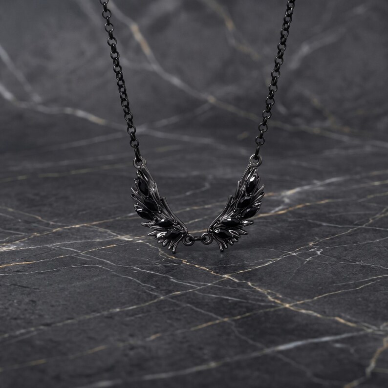Puede incluir: Un collar negro con un colgante de alas estilizadas. Las alas, con detalles texturizados, cuelgan de una fina cadena. La joya destaca sobre un fondo oscuro y veteado, resaltando su dise&ntilde;o y detalles.