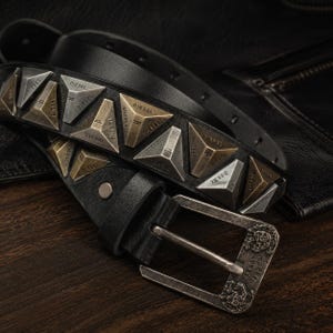 Op de afbeelding: Een zwarte riem met piramidevormige metalen studs in zilver- en bronskleuren. Elke stud is voorzien van het woord DIESEL. De riem heeft een rechthoekige metalen gesp met een gegraveerd ontwerp en een enkele lus bij de gesptong.