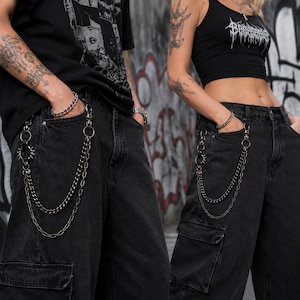 May include: Black denim cargo pants with silver chain detailing. The pants have a relaxed fit and feature multiple pockets. A silver chain hangs from the belt loops, adding a punk rock aesthetic. The pants are worn with a black top.