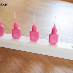 Könnte beinhalten: Vier rosa Interdentalbürsten sind auf einem weißen rechteckigen Halter angeordnet. Die Bürsten haben ein flaschenförmiges Design mit einer Bürstenspitze. Der Halter hat ein Loch auf der rechten Seite. Der Text "YBtechnology" befindet sich oben links.