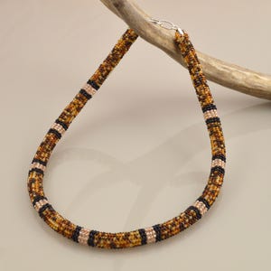 May include: A beaded necklace with a brown, tan, and black pattern. The necklace is made of small beads and has a silver clasp. The necklace is displayed on a wooden branch.