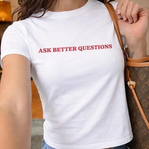 May include: A white t-shirt with the slogan "ASK BETTER QUESTIONS" printed in red. The short-sleeved top is made of a soft material. The image is a close-up of the shirt.