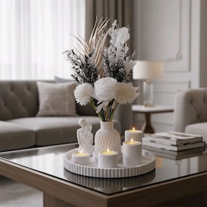 May include: A decorative living room scene featuring a white floral arrangement in a textured vase, candles, and a small sculpture on a white tray. The arrangement includes white roses, and black and gold accents. The scene is set on a glass-topped coffee table.