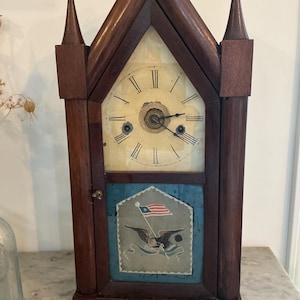 May include: A wooden antique clock with a steeple-like design. The clock face is cream-colored with black Roman numerals. Below the clock face is a blue panel with an eagle and flag design. The clock is brown.