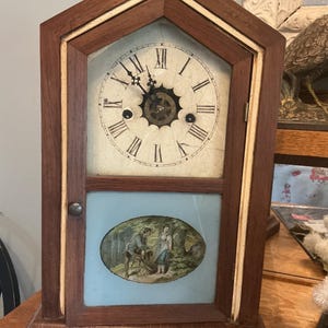 May include: Antique wooden clock with a light-colored face and Roman numerals. The clock has a dark brown frame with a light-colored trim. The bottom section features a painted oval scene of people in a forest. The clock is on a wooden surface.