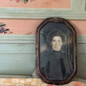 May include: An antique portrait photograph of a person with dark hair, wearing a high-collared dark garment, encased in an ornate, dark brown, octagonal wooden frame with carved floral details and a convex glass cover.