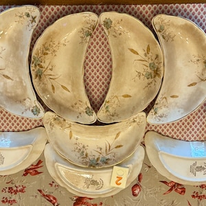 May include: A collection of antique, off-white crescent-shaped plates and serving dishes. Each piece features a floral design in muted blue and gold tones. The set includes both plates and smaller, curved dishes, ideal for appetizers or desserts.
