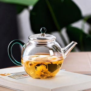 Könnte beinhalten: Eine Teekanne aus klarem Glas mit rundem Körper, kurzem Ausguss und einem dunkelgrünen Griff. Der Deckel hat einen kleinen dunkelgrünen Knauf. Im Inneren der Kanne ziehen goldgelbe Chrysanthemenblüten in einer klaren Flüssigkeit.