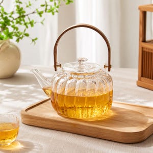 May include: A clear glass teapot with a ribbed, pumpkin-like body and a matching lid. The teapot features a curved glass spout and a tall, arched wooden handle attached with metal hardware. It is filled with a golden-coloured tea liquid.
