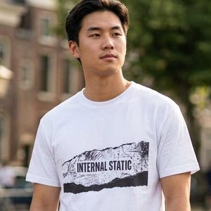 以下が含まれることがあります： 黒のダメージデザインで「INTERNAL STATIC」の文字が入った白いTシャツ。半袖でクルーネックです。ダークブルーのカーゴパンツを着用しています。背景にはヨーロッパ風の建物と橋が写っています。