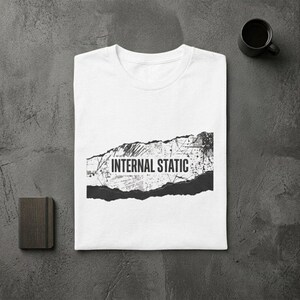 以下が含まれることがあります： 「INTERNAL STATIC」の文字が黒のダメージ加工でプリントされた白いTシャツ。Tシャツは折りたたまれ、質感のあるグレーの表面に置かれています。近くには小さなノートと黒いコーヒーマグがあります。
