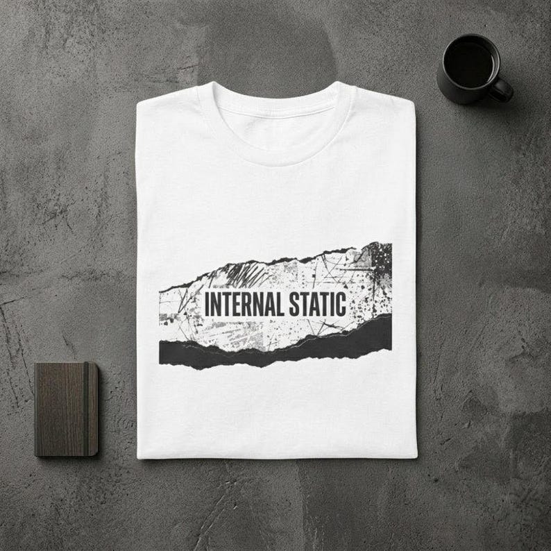 以下が含まれることがあります： 「INTERNAL STATIC」の文字が黒のダメージ加工でプリントされた白いTシャツ。Tシャツは折りたたまれ、質感のあるグレーの表面に置かれています。近くには小さなノートと黒いコーヒーマグがあります。