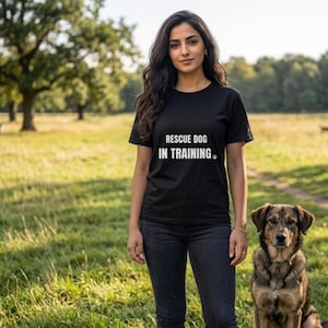 May include: A black t-shirt with the text "RESCUE DOG IN TRAINING" in white, paired with dark blue jeans. A brown and tan dog sits beside the person. The scene is set in a grassy field with trees in the background.