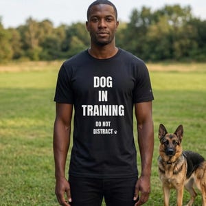 May include: A black t-shirt with the text "DOG IN TRAINING DO NOT DISTRACT" in white. A man stands in a grassy field with a German Shepherd dog. The dog is brown, black, and tan.
