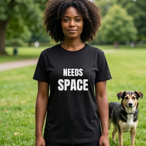 May include: A black t-shirt with the white text "NEEDS SPACE" is worn by a person standing in a park. A dog with brown, black, and white fur stands nearby. The background features green grass and trees.