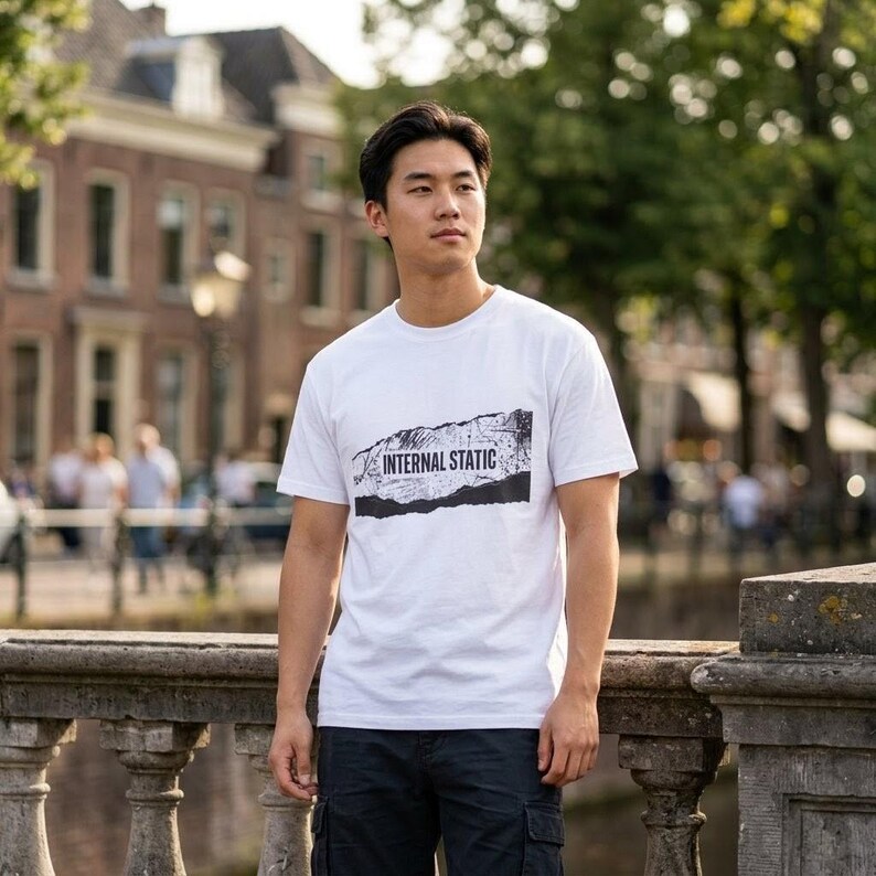 以下が含まれることがあります： 黒のダメージデザインで「INTERNAL STATIC」の文字が入った白いTシャツ。半袖でクルーネックです。ダークブルーのカーゴパンツを着用しています。背景にはヨーロッパ風の建物と橋が写っています。