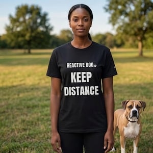 May include: A black t-shirt with the text "REACTIVE DOG KEEP DISTANCE" in white. A woman wearing the shirt stands in a grassy field with a dog. The dog is brown and white with a black collar.