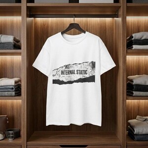 以下が含まれることがあります： 「INTERNAL STATIC」の黒い文字がプリントされた白いTシャツ。テキストは、黒い破れた紙のようなデザインの上にあります。Tシャツは、折りたたまれた服が置かれた棚のある木製のクローゼットに吊るされています。
