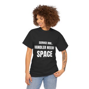 May include: Black t-shirt with the white text "SERVICE DOG. HANDLER NEEDS SPACE". The shirt is worn by a person with curly brown hair and blue jeans.