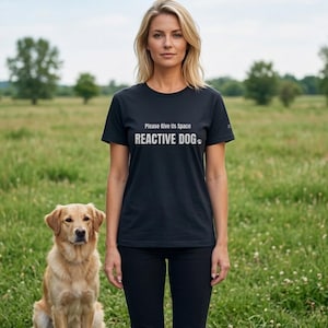 May include: Black t-shirt with the text "Please Give Us Space REACTIVE DOG" in white. A golden retriever sits in the grass next to a person wearing the shirt. The background is a green field with trees.