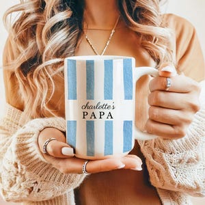 May include: White ceramic mug with vertical light blue stripes. The mug features the text "Charlotte's PAPA" in black script and block letters. The mug has a white handle and is held by a person.