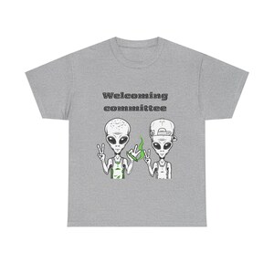 Op de afbeelding: Een grijs t-shirt met de tekst "Welcoming Committee" boven een afbeelding van twee cartoonaliens. De aliens hebben grote ogen en geven vredestekens. Een alien houdt een groen object vast.