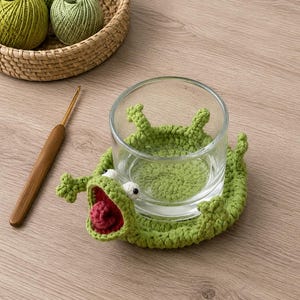Peut inclure: Un dessous de verre en crochet vert clair en forme de grenouille avec une bouche grande ouverte, une langue rouge et des yeux blancs, entourant un verre transparent. Un crochet en bois et un panier contenant trois pelotes de laine verte.