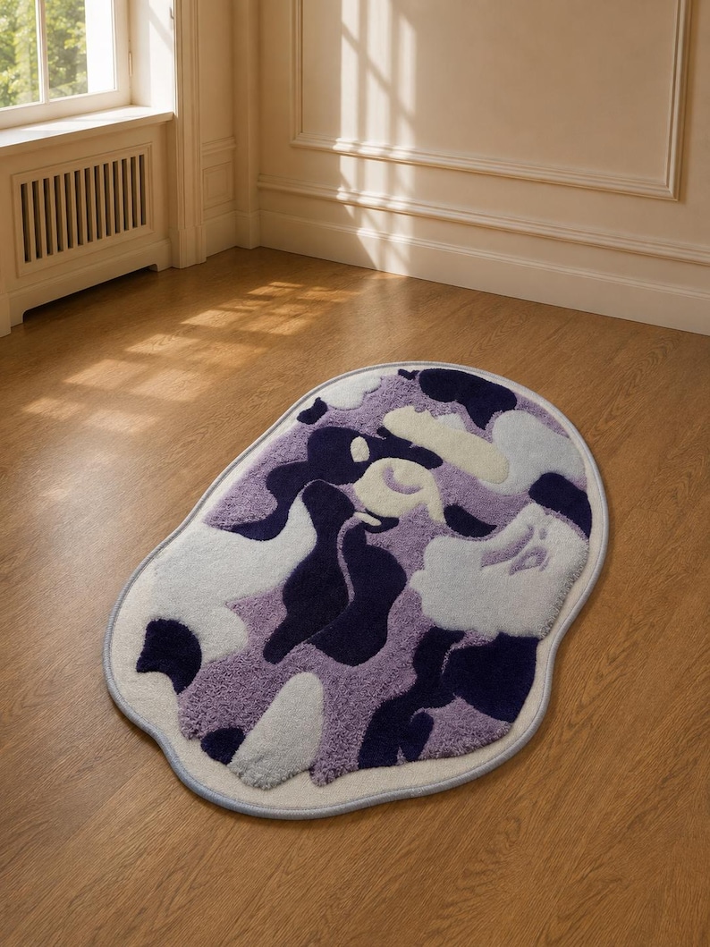 May include: An oval-shaped area rug featuring an abstract camouflage pattern in shades of deep navy blue, light lavender, and soft white. The rug has a textured, tufted pile surface and is finished with a light blue bound edge trim.