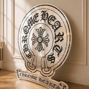 Könnte beinhalten: Ein großes, kreisförmiges, reflektierendes Schild mit dem schwarzen Schriftzug CHROME HEARTS in gotischer Schrift, der kreisförmig um ein zentrales schwarzes Kreuzemblem angeordnet ist. Darunter zeigt eine Basis den Text CHROME HEARTS.