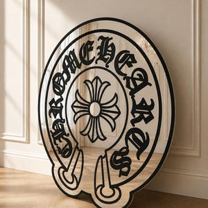 Könnte beinhalten: Ein großer runder Spiegel mit einem schwarzen Rahmen und einem schwarzen grafischen Design auf der Glasoberfläche. Das Motiv zeigt den Schriftzug CHROME HEARTS in gotischer Schrift in Hufeisenform um ein zentrales vierzackiges Kreuz.