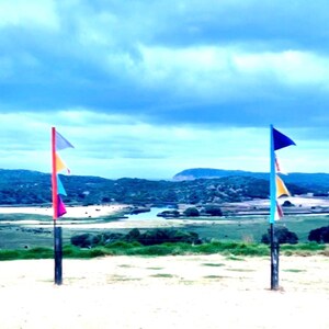 May include: Two flag poles with colorful triangular flags stand in the foreground against a scenic landscape. The flags are in shades of pink, orange, blue, and gray. The background features a river, green hills, and a cloudy blue sky.