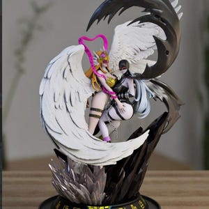 Può includere: Statuetta dettagliata con due personaggi in stile anime con grandi ali. Un personaggio ha i capelli biondi e un abito dorato, l'altro ha i capelli neri e un abito nero. La base è nera con testo giapponese e una struttura cristallina.