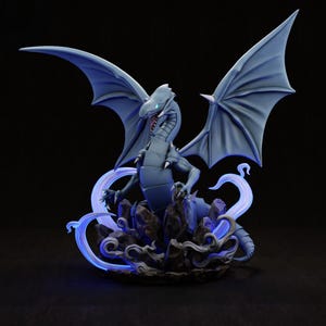 Può includere: Statuetta dettagliata di un drago blu con ali spiegate, appollaiato su una base rocciosa. Il drago ha artigli affilati, un'espressione minacciosa e accenti blu luminosi. Le ali sono spalancate e la base ha un'aura blu brillante.