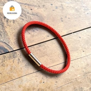 May include: A red rope bracelet with a gold-colored clasp. The bracelet is made of a twisted, textured rope material. The clasp is a cylindrical shape. The bracelet is shown on a wooden surface.