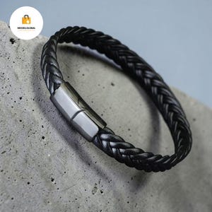 May include: Black braided leather bracelet with a silver clasp. The bracelet is made of woven leather and has a simple design. The rectangular clasp appears to be metal. The bracelet is displayed on a grey surface.