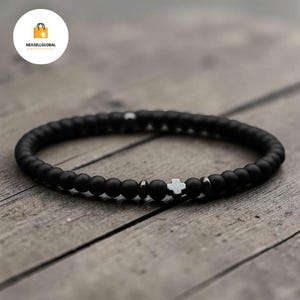 May include: Black beaded bracelet featuring a white cross charm and silver details. The bracelet is composed of small, round, matte black beads. The cross charm is positioned centrally.